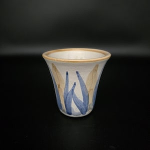Può includere: Un vaso in ceramica con esterno color crema, decorato con disegni astratti blu e beige a forma di foglia. Il bordo è color beige chiaro. Il vaso ha una forma leggermente svasata e poggia su una superficie nera.