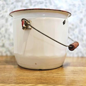 May include: Cream-colored enamel bucket with a red rim and a metal handle with a wooden grip. The bucket has a slightly aged appearance, suggesting vintage or antique status. The handle is attached with a red-painted hinge.