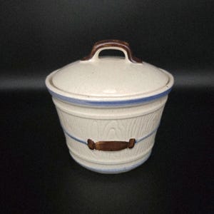 May include: A cream-colored ceramic container with a lid. The container has a wood-grain texture, blue accents, and brown handles. The lid has a brown handle. The container is approximately 6 inches tall.
