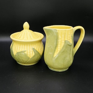 Puede incluir: Un azucarero de cerámica con tapa y una jarra para crema, ambos con diseño de mazorca de maíz. Las piezas son amarillas con detalles verdes. El azucarero tiene un pomo en la parte superior. La jarra tiene asa y pico.