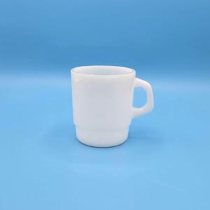 Könnte beinhalten: Eine weiße Keramiktasse mit Henkel. Die Tasse ist aus Milchglas und hat ein schlichtes Design.