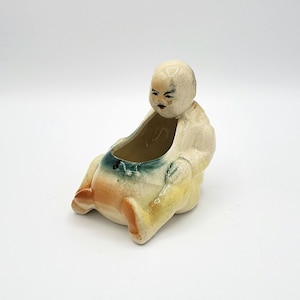 May include: A ceramic figurine of a seated man with a green and yellow glaze. The figurine has a hollow center that can be used as a small dish or planter.