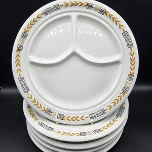 May include: A stack of white ceramic divided plates featuring a three-section design. The rim of each plate is decorated with a repeating pattern of black geometric square motifs and gold-colored laurel leaf sprigs arranged in a circular border.