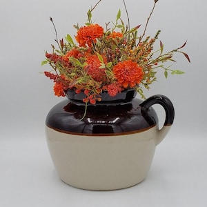 Puede incluir: Un tarro de cerámica marrón y blanco con asa, lleno de un arreglo floral de flores naranjas y verdes.