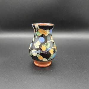 May include: A small, handcrafted ceramic vase with a black base and colorful polka dots in blue, yellow, and white. The vase has a flared rim and a brown base, perfect for displaying small flowers or decorative items.