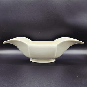 May include: Cream-colored, vintage-style ceramic bowl with a unique, symmetrical design. The bowl has a wide, open center and flared, wing-like edges. It sits on a small, circular base, and is set against a dark background.