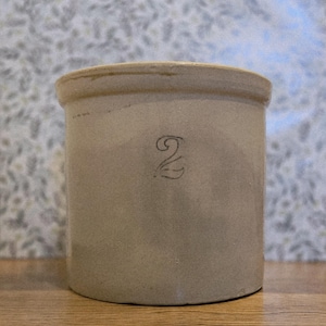 Puede incluir: Un tarro cilíndrico de gres de color beige claro. El número "2" está estampado en negro en la parte delantera. El tarro tiene un borde redondeado y descansa sobre una superficie de madera.