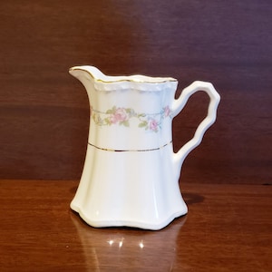 May include: A white ceramic creamer with a gold trim and a floral design. The creamer has a handle and a spout.