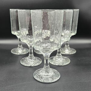 May include: A set of six clear, textured glass goblets. Each goblet has a square bowl with vertical ridges and a textured stem and base. The glasses are arranged in a semi-circle.