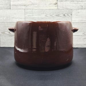 Puede incluir: Una olla de cocina de cerámica marrón oscuro con dos asas pequeñas en los lados. La olla tiene un acabado brillante y una forma redondeada. El fondo es una pared de paneles de madera gris claro y blanco.