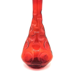 May include: A red glass vase with a distinctive shape and a series of raised circular designs. The vase has a narrow neck and a wider, rounded body. The colour is a deep, rich red.