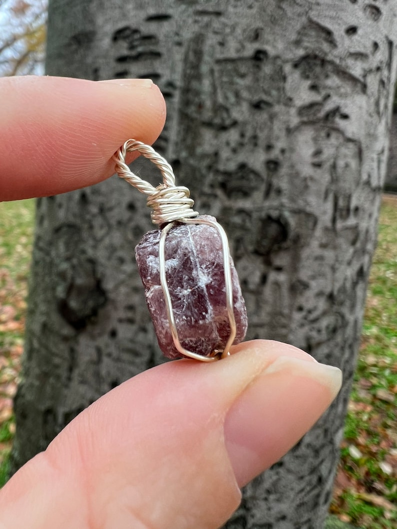 May include: A purple and silver wire-wrapped crystal pendant. The crystal is a natural, rough-cut stone with a deep purple hue. The wire wrapping is silver and adds a delicate touch to the pendant.