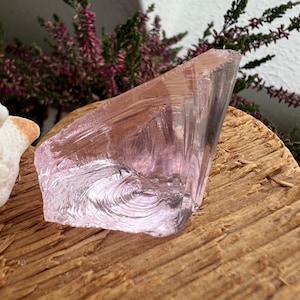 May include: A translucent, light pink crystal with a rough, textured surface. The crystal is irregularly shaped, with a sharp, angled edge. The crystal sits on a piece of weathered wood, with sprigs of purple flowers in the background.
