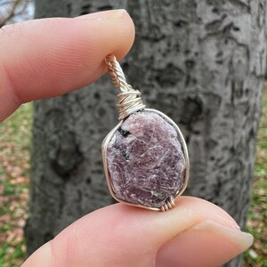 May include: A silver wire-wrapped pendant featuring a raw, pink and purple gemstone with black inclusions.