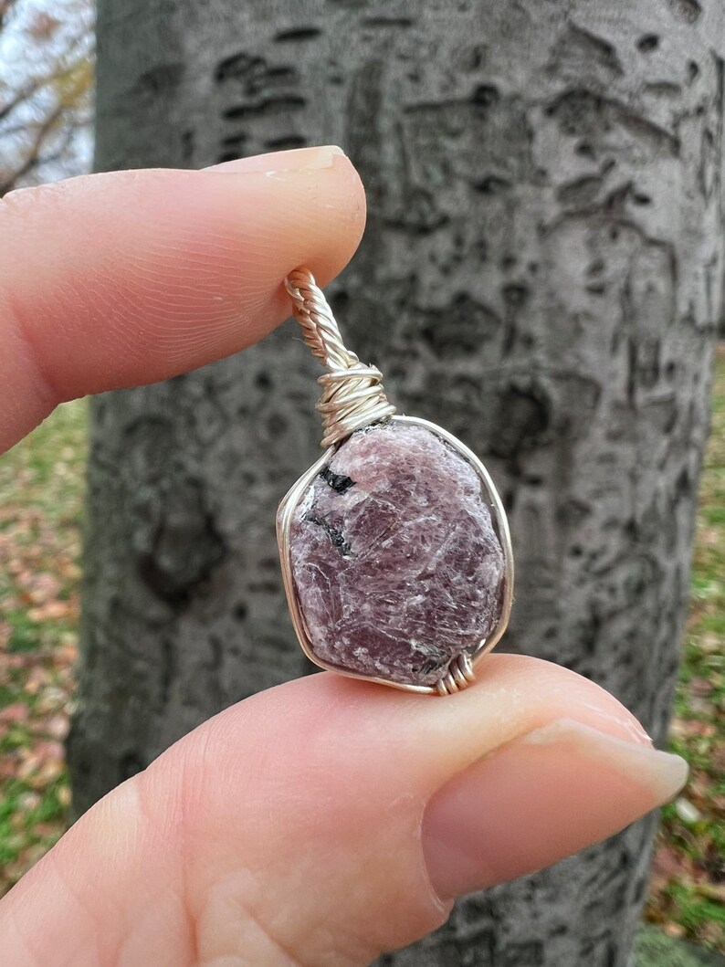 May include: A silver wire-wrapped pendant featuring a raw, pink and purple gemstone with black inclusions.