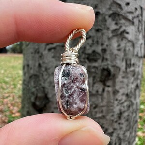 May include: A silver wire-wrapped pendant featuring a raw, pink and grey gemstone. The pendant is held in a hand against a tree trunk background.