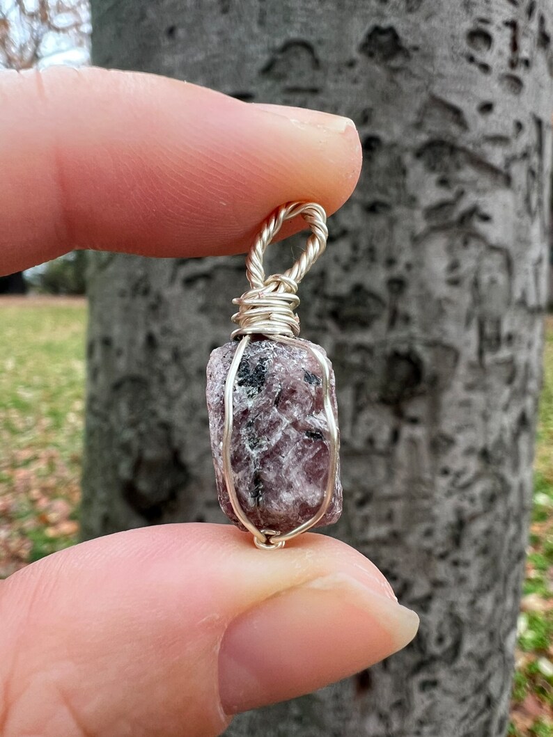 May include: A silver wire-wrapped pendant featuring a raw, pink and grey gemstone. The pendant is held in a hand against a tree trunk background.