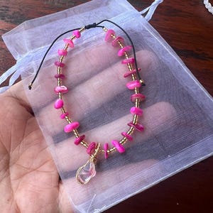 Könnte beinhalten: Ein Armband aus rosa Edelsteinen mit goldfarbenen Perlen und einem klaren, tropfenförmigen Anhänger. Das Armband befindet sich an einem schwarzen Kordel und wird in einem weißen Netzbeutel präsentiert. Es ist ein Schmuckstück.