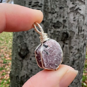 May include: A pink and purple gemstone pendant wrapped in silver wire. The gemstone has a rough, natural texture.