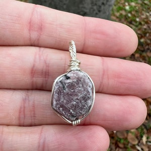 May include: A silver wire wrapped pendant featuring a raw, pink, and gray gemstone. The pendant is oval shaped and has a rough, natural texture.