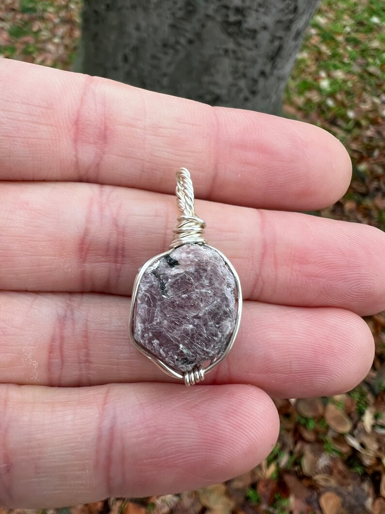 May include: A silver wire wrapped pendant featuring a raw, pink, and gray gemstone. The pendant is oval shaped and has a rough, natural texture.