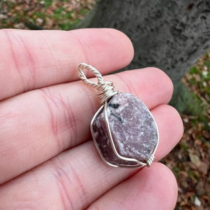 May include: A pink and grey gemstone pendant wrapped in silver wire. The pendant is a raw, uncut stone with a rough, natural texture.