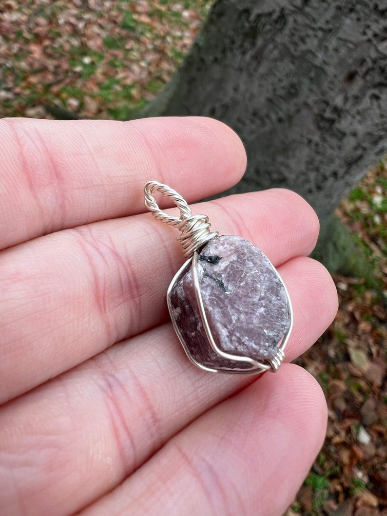 May include: A pink and grey gemstone pendant wrapped in silver wire. The pendant is a raw, uncut stone with a rough, natural texture.