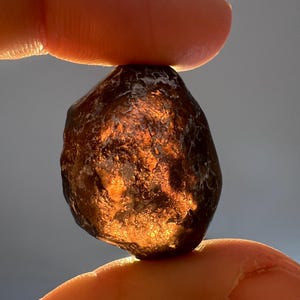 Peut inclure: Une pierre précieuse brun foncé de forme irrégulière tenue entre deux doigts. La surface de la pierre reflète la lumière, créant un effet scintillant. La pierre précieuse semble être un minéral naturel.