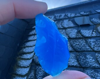 May include: A vibrant, translucent blue glass shard is held between two fingers against a backdrop of a cloudy sky and a dark roof. The shard has irregular edges and a smooth, polished surface, catching the light.