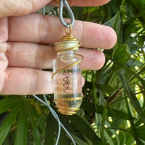 May include: A gold-wrapped glass vial necklace with a clear liquid and a gold flower of life symbol inside. The necklace is on a gray cord.
