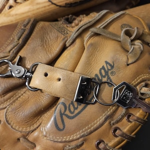 Op de afbeelding: Een bruine leren honkbalhandschoen met het woord "Rangers" erop gestikt. Een sleutelhanger met een leren band en een zilveren sleutel is aan de handschoen bevestigd.