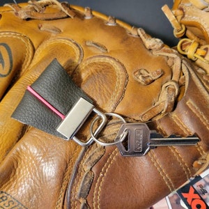 Könnte beinhalten: Ein brauner Leder-Baseballhandschuh mit einem schwarzen Leder-Schlüsselanhänger. Der Schlüsselanhänger hat eine silberne Metallschlaufe und einen roten Streifen. Ein silberner Schlüssel ist am Schlüsselanhänger befestigt.
