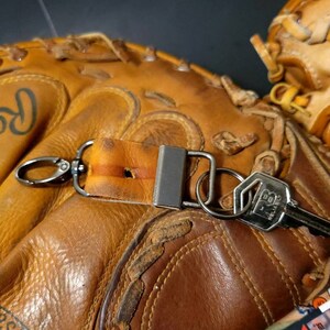 Könnte beinhalten: Ein brauner Leder-Baseballhandschuh mit einem Schlüsselanhänger. Der Schlüsselanhänger hat einen Lederriemen, einen silbernen Metallring und einen silbernen Schlüssel mit dem Text "BB" darauf.