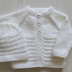 May include: A white knitted baby cardigan and matching hat set. The cardigan has a button closure and a delicate stitch pattern. The hat is a simple beanie style.