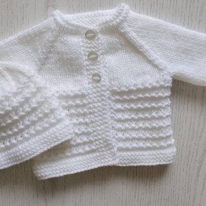 May include: A white knitted baby cardigan and matching hat set. The cardigan has a button closure and the hat is a simple beanie style.