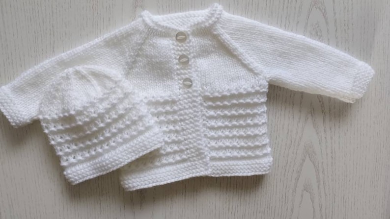 May include: A white knitted baby cardigan and matching hat set. The cardigan has a button closure and the hat is a simple beanie style.