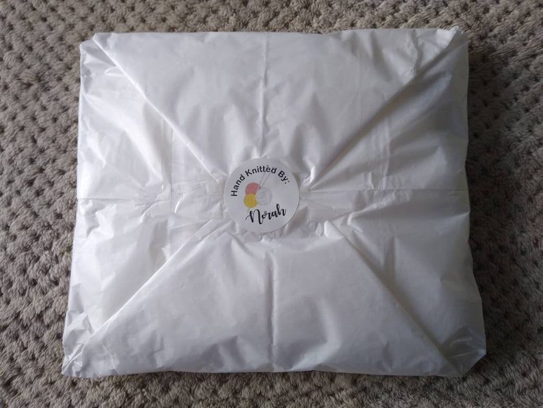 May include: White tissue paper packaging with a round sticker that says "Hand Knitted By: Norah" with a colorful yarn ball and knitting needles graphic.