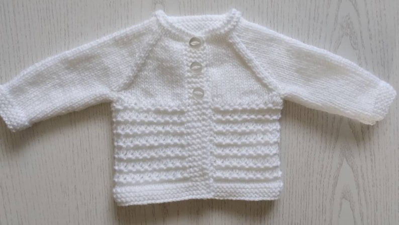 May include: White knitted baby cardigan with a button closure. The cardigan has a textured pattern and is made of soft yarn.