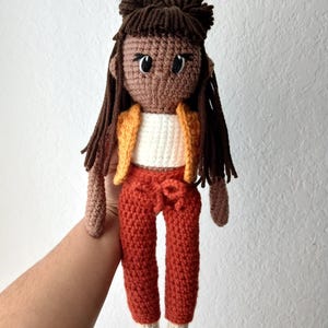 Puede incluir: Muñeca de ganchillo hecha a mano con piel morena, cabello castaño oscuro y ojos negros grandes. La muñeca viste un top blanco, una chaqueta naranja y pantalones color óxido. Los pies de la muñeca son de color crema.
