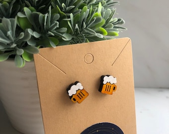 May include: A pair of wooden beer mug earrings. The mugs are painted brown with white foam on top. The earrings are on a brown card with a blue circle that says "1150 Creative".