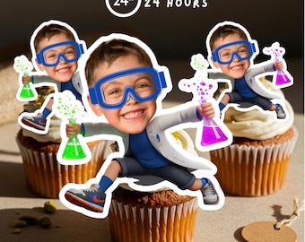 May include: Three cupcakes decorated with cartoon images of a boy in blue safety goggles and a lab coat, holding a flask with colourful bubbling liquid. The text "READY IN 24 HOURS" is in a circle above the cupcakes.