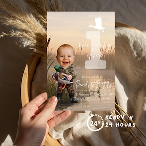 May include: A birthday invitation featuring a baby in camouflage holding a toy duck. The text "one lucky duck" is above the baby. The invitation includes event details and a "Ready in 24 hours" graphic.