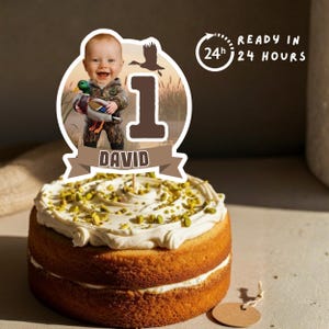 May include: A round cake with white frosting and green sprinkles, topped with a cake topper featuring a baby holding a duck, the number 1, and the name "DAVID". The topper also includes the text "READY IN 24 HOURS".