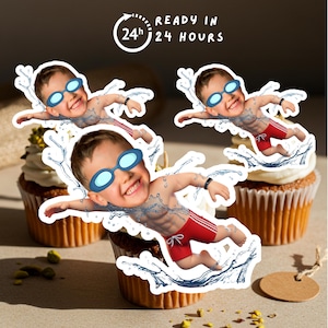 May include: Three cupcakes with white frosting are decorated with images of a child swimming. The child is wearing goggles and red swim trunks. The image includes the text "READY IN 24 HOURS".