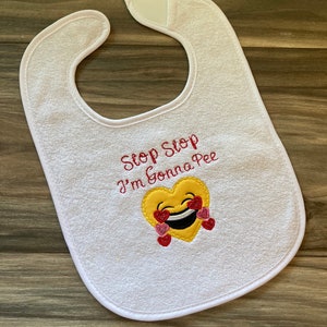 May include: White baby bib with red embroidered text that says "Stop Stop I'm Gonna Pee" and a yellow heart emoji with red hearts around it.