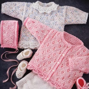 Può includere: Una collezione di capi per neonati lavorati a maglia a mano che include un maglione bianco maculato con colletto in pizzo, un cardigan rosa abbottonato, pantaloncini bianchi, una cuffietta rosa con nastri e due paia di scarpine bianche e rosa.