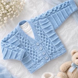 May include: A light blue knitted cardigan featuring a V-neckline, four matching buttons, and a textured cable knit pattern on the front and sleeves. The garment is layered over a plain white shirt and is accompanied by white booties and a teddy bear.