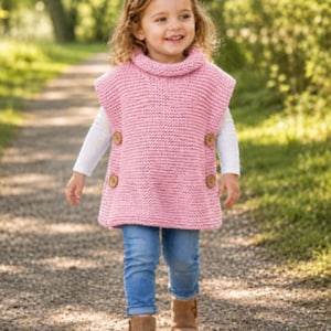 May include: A young child wearing a pink knitted poncho with a turtleneck collar and wooden buttons. The child is also wearing blue jeans, a white long-sleeved shirt, and brown boots. The child is walking on a gravel path.