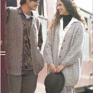 Op de afbeelding: Een stel draagt gebreide kleding. De man draagt een bruin gebreid vest over een shirt met patroon en een bruine broek. De vrouw draagt een lichtgrijze cardigan over een wit shirt en een rok met patroon, met een zwarte hoed in haar handen. De kleding lijkt handgemaakt.