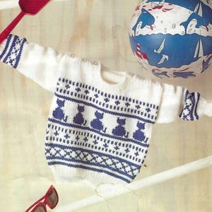 May include: A white long-sleeve sweater featuring a blue knit pattern of sitting cats and geometric shapes. The sweater has ribbed cuffs and a crew neckline. Accompanying items include a red plastic shovel, red sunglasses, and a blue beach ball.